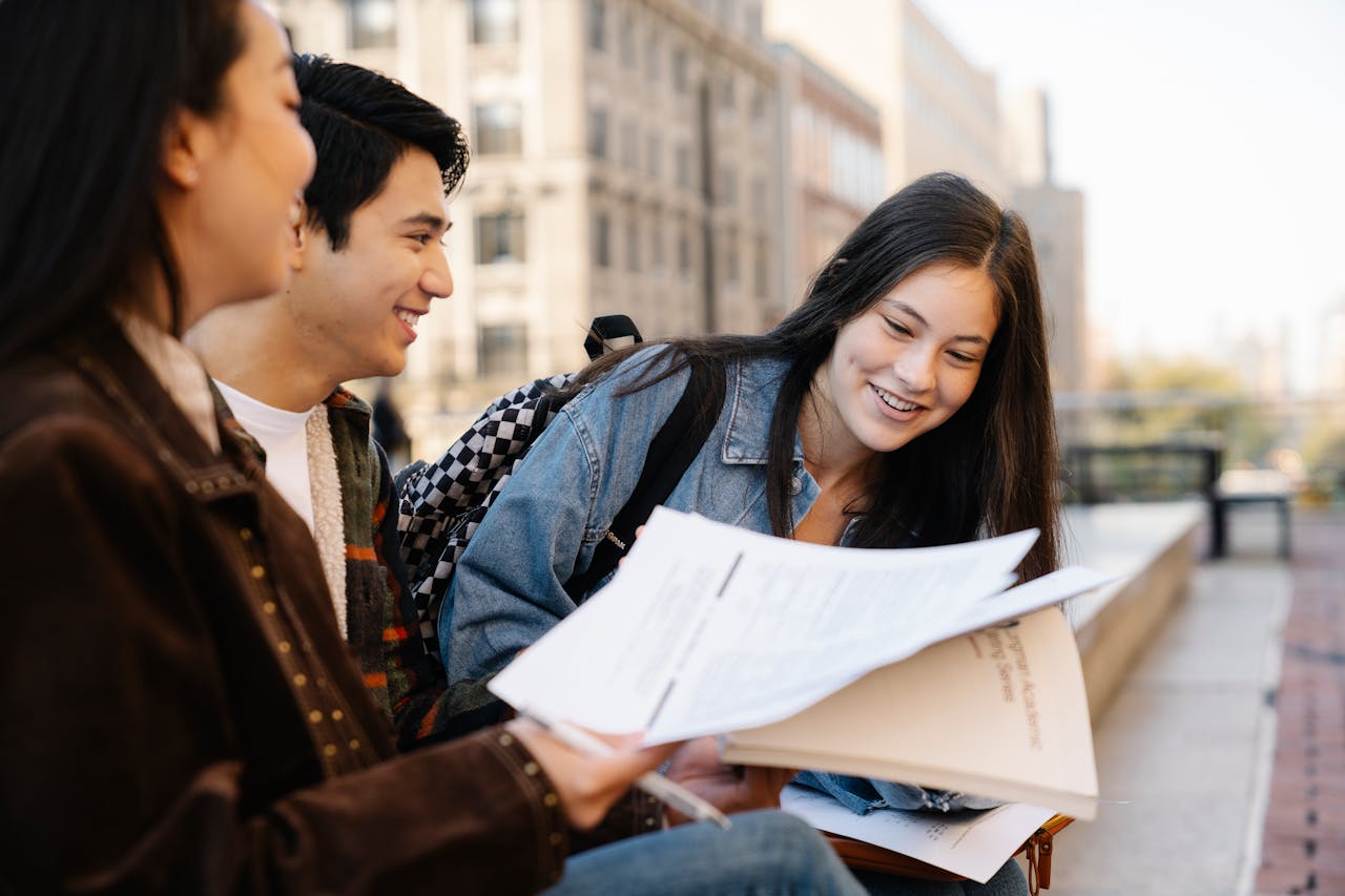 Teen students engage in outdoor study session, sharing documents and smiling.